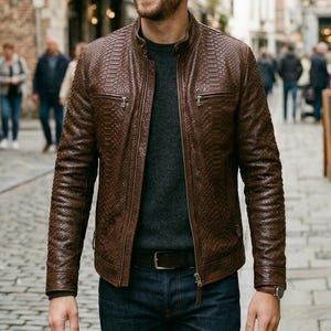 May include: A brown leather jacket with a textured pattern, worn over a gray sweater and dark blue jeans. The jacket has a zipper closure and a classic collar. The person is walking on a cobblestone street.
