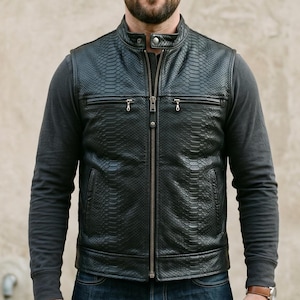 May include: A black leather vest with a snakeskin pattern, featuring a zip-up front and a stand-up collar. The vest is worn over a gray long-sleeved shirt and paired with blue jeans. A watch is visible on the wrist.
