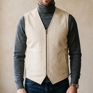 May include: A cream-colored vest with a reptile-like texture and a full-length zipper. The vest is worn over a gray turtleneck sweater and blue jeans. The person is wearing a watch with a brown leather strap.