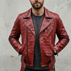 Könnte beinhalten: Rote Lederjacke mit strukturiertem, krokodilähnlichem Muster. Die Jacke hat einen klassischen Biker-Stil mit asymmetrischem Reißverschluss, einer kleinen Tasche und einem Gürtel. Das Model trägt ein graues T-Shirt darunter.