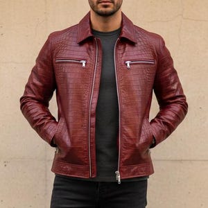 Könnte beinhalten: Burgunderfarbene Lederjacke mit strukturiertem Muster, Reißverschluss und zwei Vordertaschen. Die Jacke wird mit einem dunkelgrauen Hemd, schwarzen Jeans und schwarzen Stiefeln kombiniert. Das Outfit befindet sich vor einem neutralen Hintergrund.