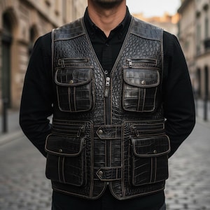 Op de afbeelding: Een donkerbruin leren vest met meerdere zakken en een ritssluiting. Het vest heeft een textuur met krokodillenpatroon en is afgewerkt met lichtbruine stiksels. Het vest wordt over een zwart overhemd gedragen.