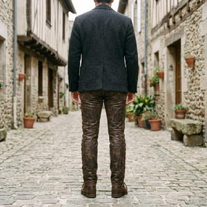 May include: A charcoal gray blazer with a textured finish and a single back vent, paired with distressed brown leather trousers that feature a worn, vintage appearance and brown leather boots, viewed from the back in a narrow stone alleyway.