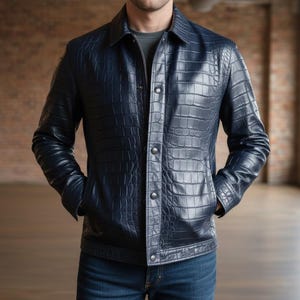 May include: A navy blue leather jacket with a crocodile skin texture. The jacket has a button-up front, two front pockets, and a collared neckline. The jacket is worn over a gray shirt and blue jeans.
