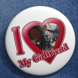 Pode incluir: Um botão branco com o texto "I My Girlfriend" em vermelho, com uma moldura em forma de coração vermelho contendo uma imagem de uma pessoa com um cocar de penas. O botão está preso a uma jaqueta jeans.