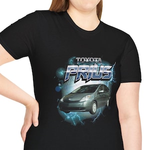 Peut inclure: T-shirt noir avec un motif d'une voiture Toyota Prius et le texte "TOYOTA PRIUS". La voiture est argentée, sur fond de motifs d'éclairs bleus et blancs. Le t-shirt est porté par une personne.