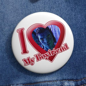 Puede incluir: Un botón blanco con el texto "I My Boyfriend" en rojo, con un marco en forma de corazón alrededor de un personaje azul de la película Avatar. El botón está prendido a una chaqueta vaquera.