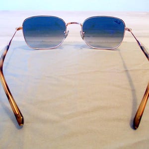 Pu&ograve; includere: Occhiali da sole quadrati con lenti blu sfumate e montatura color oro. Le aste presentano un motivo tartarugato. Il logo Ray-Ban &egrave; visibile sulla lente superiore destra. Gli occhiali da sole poggiano su un panno grigio chiaro.