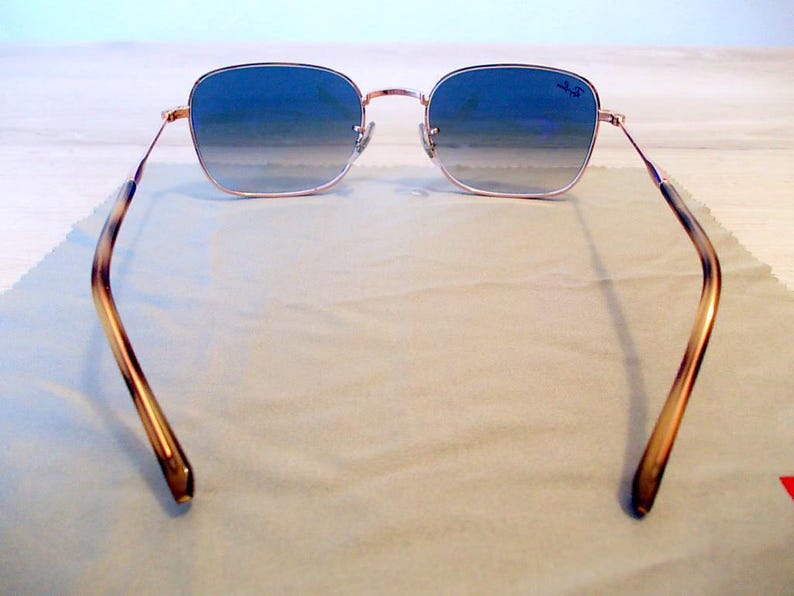 Pu&ograve; includere: Occhiali da sole quadrati con lenti blu sfumate e montatura color oro. Le aste presentano un motivo tartarugato. Il logo Ray-Ban &egrave; visibile sulla lente superiore destra. Gli occhiali da sole poggiano su un panno grigio chiaro.
