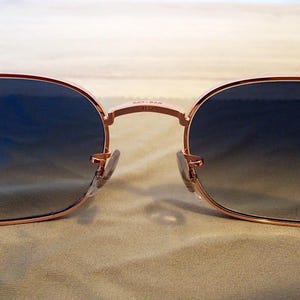 Pu&ograve; includere: Primo piano di occhiali da sole con montatura color oro rosa e lenti sfumate blu. Il logo Ray-Ban &egrave; visibile sul ponte. Gli occhiali da sole hanno una forma classica e arrotondata, con naselli.