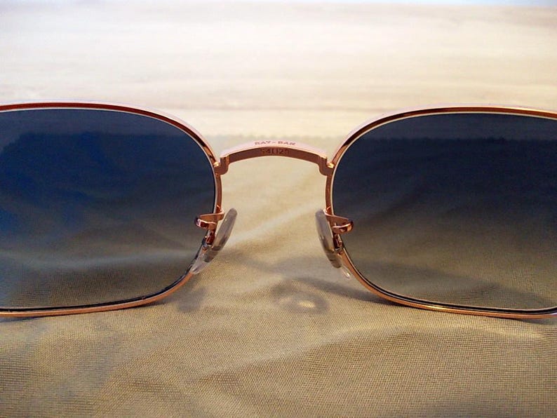 Pu&ograve; includere: Primo piano di occhiali da sole con montatura color oro rosa e lenti sfumate blu. Il logo Ray-Ban &egrave; visibile sul ponte. Gli occhiali da sole hanno una forma classica e arrotondata, con naselli.