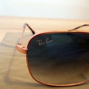 Pu&ograve; includere: Primo piano di occhiali da sole Ray-Ban con montatura color oro e lenti sfumate. Il logo Ray-Ban &egrave; visibile sulla lente. Gli occhiali da sole sono appoggiati su un panno verde chiaro.