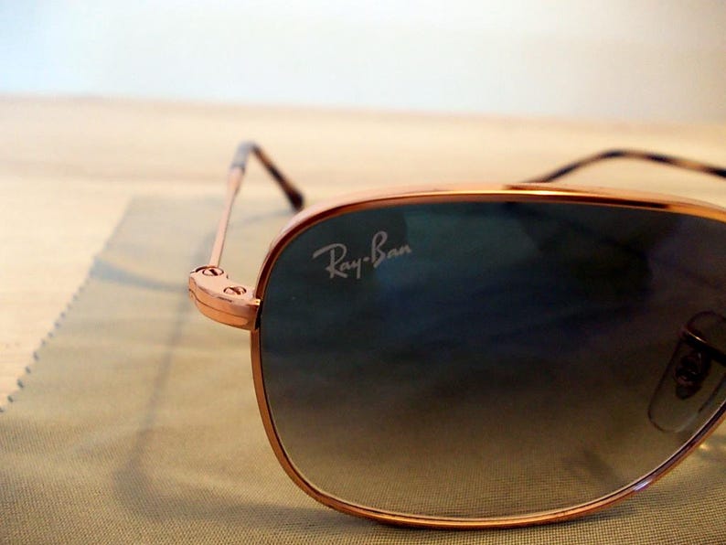 Pu&ograve; includere: Primo piano di occhiali da sole Ray-Ban con montatura color oro e lenti sfumate. Il logo Ray-Ban &egrave; visibile sulla lente. Gli occhiali da sole sono appoggiati su un panno verde chiaro.