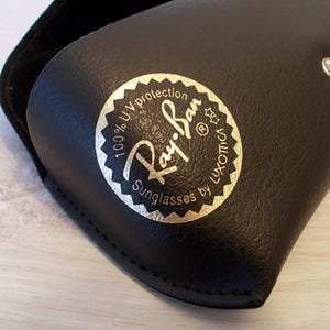 Pu&ograve; includere: Custodia per occhiali da sole in pelle nera con logo Ray-Ban dorato e bianco. La custodia ha una chiusura a scatto e una morbida fodera interna. Il logo recita "100% UV protection" e "Sunglasses by Luxottica."