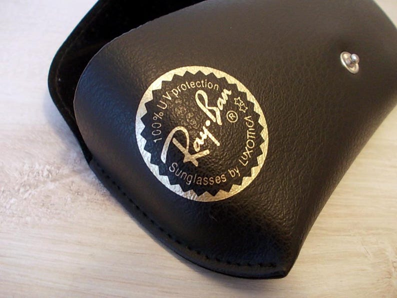 Pu&ograve; includere: Custodia per occhiali da sole in pelle nera con logo Ray-Ban dorato e bianco. La custodia ha una chiusura a scatto e una morbida fodera interna. Il logo recita "100% UV protection" e "Sunglasses by Luxottica."