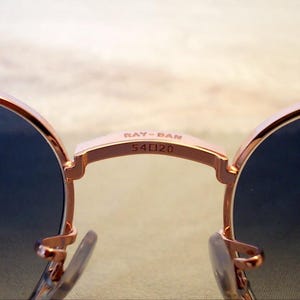 Pu&ograve; includere: Primo piano di occhiali da sole Ray-Ban con montatura oro rosa e lenti blu scuro. Il ponte degli occhiali mostra il nome del marchio e le informazioni sulla taglia: "RAY-BAN 54口20". Gli occhiali da sole hanno una forma classica e arrotondata.