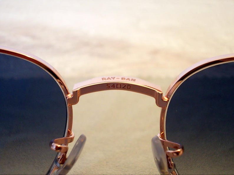 Pu&ograve; includere: Primo piano di occhiali da sole Ray-Ban con montatura oro rosa e lenti blu scuro. Il ponte degli occhiali mostra il nome del marchio e le informazioni sulla taglia: "RAY-BAN 54口20". Gli occhiali da sole hanno una forma classica e arrotondata.