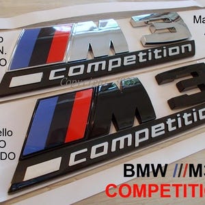 Puede incluir: Dos emblemas BMW M3 Competition. Uno es plateado con rayas azules, rojas y negras, y el otro es negro con las mismas rayas. El texto "COMPETITION" es blanco. El texto "BMW ///M3 COMPETITION" está en la parte inferior.