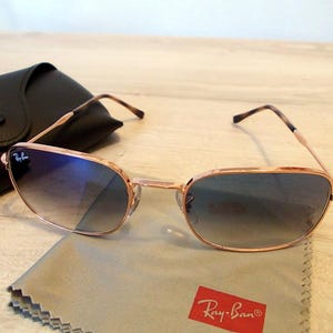 Pu&ograve; includere: Occhiali da sole color oro rosa con lenti sfumate blu, custodia in pelle nera e panno per la pulizia con logo Ray-Ban. Gli occhiali hanno una forma rettangolare e un design classico.