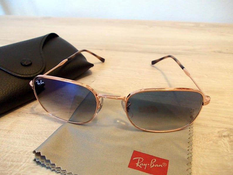 Pu&ograve; includere: Occhiali da sole color oro rosa con lenti sfumate blu, custodia in pelle nera e panno per la pulizia con logo Ray-Ban. Gli occhiali hanno una forma rettangolare e un design classico.