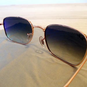 Pu&ograve; includere: Occhiali da sole con montatura color oro rosa e lenti blu sfumate. Le montature quadrate e le aste sottili hanno un design classico. Le lenti passano dal blu scuro al blu chiaro. Il logo Ray-Ban &egrave; visibile sulla lente.