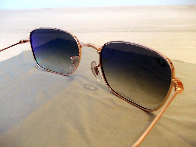 Pu&ograve; includere: Occhiali da sole con montatura color oro rosa e lenti blu sfumate. Le montature quadrate e le aste sottili hanno un design classico. Le lenti passano dal blu scuro al blu chiaro. Il logo Ray-Ban &egrave; visibile sulla lente.