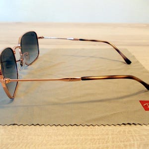 Pu&ograve; includere: Occhiali da sole color oro rosa con montatura quadrata e lenti sfumate. Le aste presentano un motivo tartarugato marrone. Gli occhiali da sole poggiano su un panno per la pulizia grigio con logo Ray-Ban rosso.