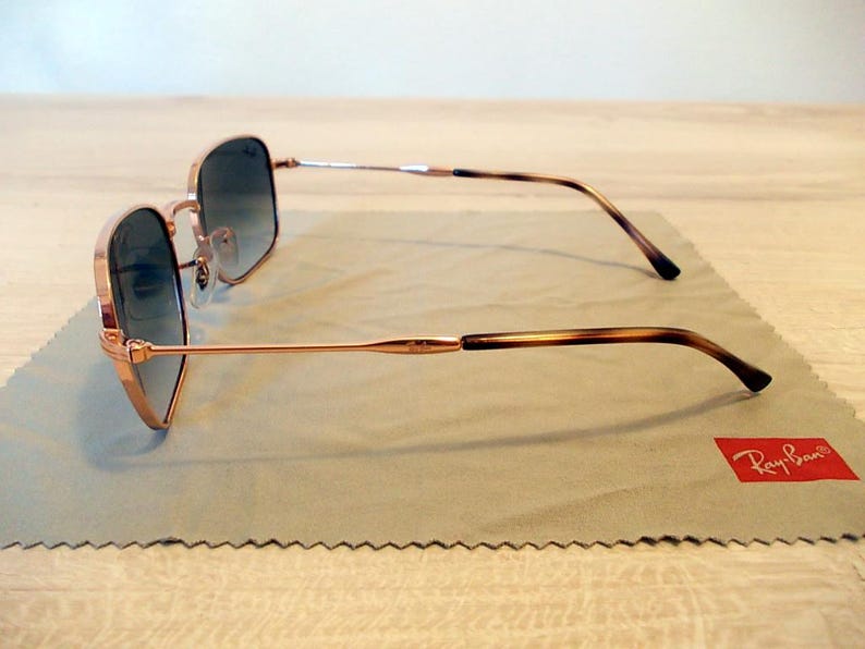 Pu&ograve; includere: Occhiali da sole color oro rosa con montatura quadrata e lenti sfumate. Le aste presentano un motivo tartarugato marrone. Gli occhiali da sole poggiano su un panno per la pulizia grigio con logo Ray-Ban rosso.