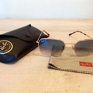 Pu&ograve; includere: Occhiali da sole con montatura dorata e lenti sfumate blu, accompagnati da un astuccio in pelle nera e un panno per la pulizia grigio. Il panno e l'astuccio riportano il logo Ray-Ban. L'insieme &egrave; su una superficie chiara.