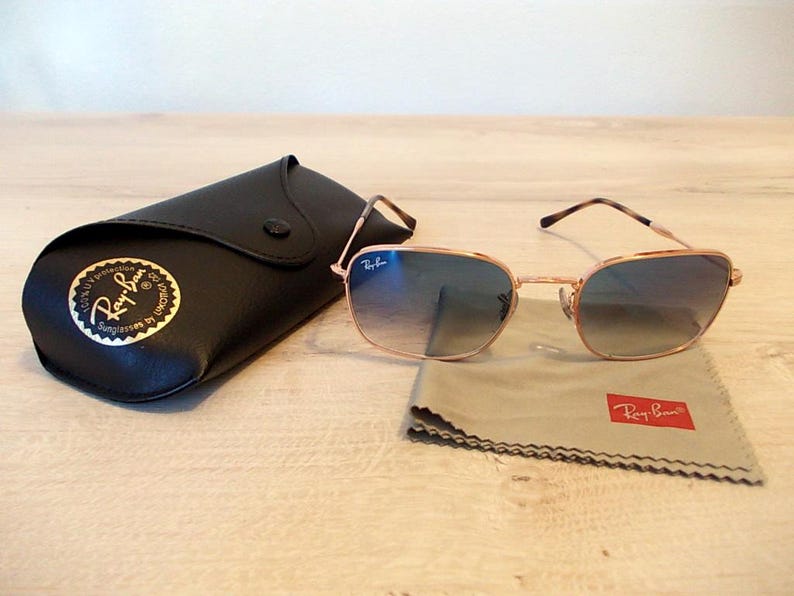 Pu&ograve; includere: Occhiali da sole con montatura dorata e lenti sfumate blu, accompagnati da un astuccio in pelle nera e un panno per la pulizia grigio. Il panno e l'astuccio riportano il logo Ray-Ban. L'insieme &egrave; su una superficie chiara.