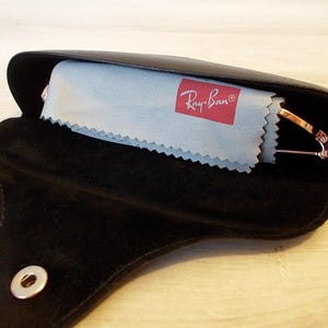 Pu&ograve; includere: Custodia per occhiali in pelle nera con chiusura a scatto, aperta per rivelare un panno per la pulizia azzurro con il logo Ray-Ban e un paio di occhiali con montatura dorata. La custodia &egrave; aperta su una superficie di legno chiaro.