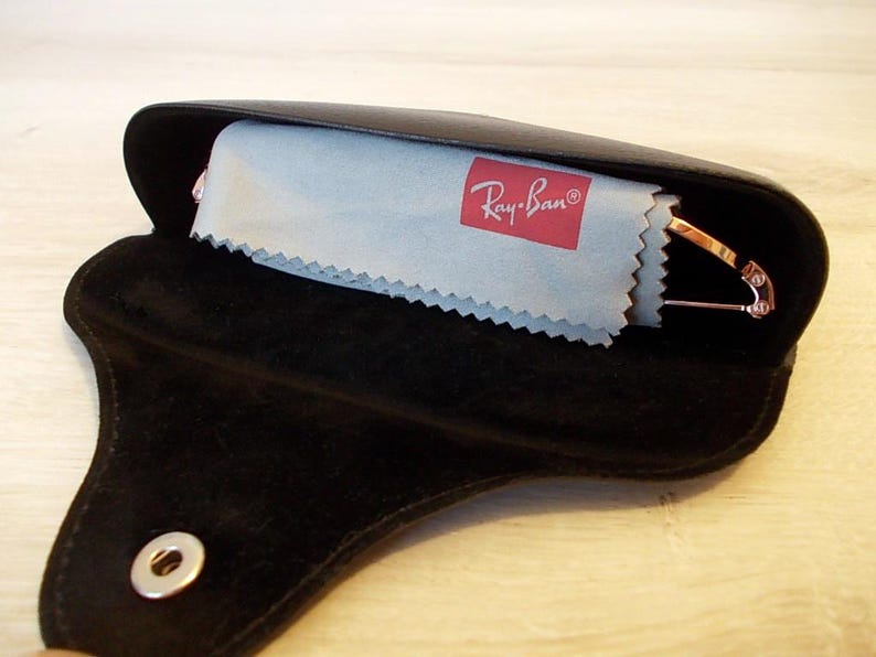 Pu&ograve; includere: Custodia per occhiali in pelle nera con chiusura a scatto, aperta per rivelare un panno per la pulizia azzurro con il logo Ray-Ban e un paio di occhiali con montatura dorata. La custodia &egrave; aperta su una superficie di legno chiaro.