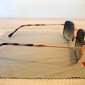 Pu&ograve; includere: Occhiali da sole dorati con lenti sfumate appoggiati su un panno per la pulizia verde chiaro. Le aste hanno un motivo tartarugato. Il logo Ray-Ban &egrave; visibile. Design classico ed elegante.