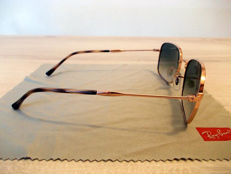 Pu&ograve; includere: Occhiali da sole dorati con lenti sfumate appoggiati su un panno per la pulizia verde chiaro. Le aste hanno un motivo tartarugato. Il logo Ray-Ban &egrave; visibile. Design classico ed elegante.