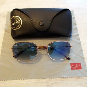 Pu&ograve; includere: Occhiali da sole quadrati con montatura dorata e lenti sfumate blu. Sono appoggiati su un panno per la pulizia grigio, accanto a una custodia in pelle nera. Il logo Ray-Ban &egrave; visibile sugli occhiali, sulla custodia e sul panno.