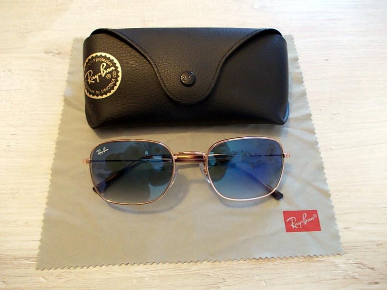 Pu&ograve; includere: Occhiali da sole quadrati con montatura dorata e lenti sfumate blu. Sono appoggiati su un panno per la pulizia grigio, accanto a una custodia in pelle nera. Il logo Ray-Ban &egrave; visibile sugli occhiali, sulla custodia e sul panno.