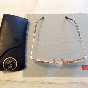 Pu&ograve; includere: Occhiali da sole con montatura dorata e lenti scure appoggiati su un panno per la pulizia grigio. A sinistra, una custodia in pelle nera con bottone e logo dorato. Il logo Ray-Ban &egrave; visibile.