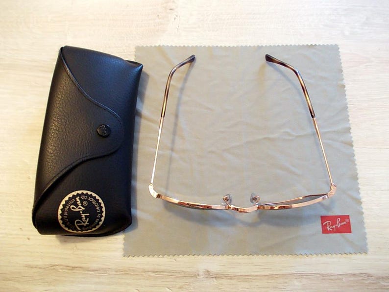 Pu&ograve; includere: Occhiali da sole con montatura dorata e lenti scure appoggiati su un panno per la pulizia grigio. A sinistra, una custodia in pelle nera con bottone e logo dorato. Il logo Ray-Ban &egrave; visibile.