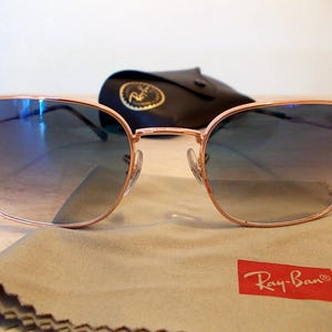Pu&ograve; includere: Occhiali da sole con montatura color oro rosa e lenti blu sfumate. Il logo Ray-Ban &egrave; visibile sulla lente superiore sinistra e su un rettangolo rosso sul panno per la pulizia. Sullo sfondo, una custodia nera con un'emblema dorato.