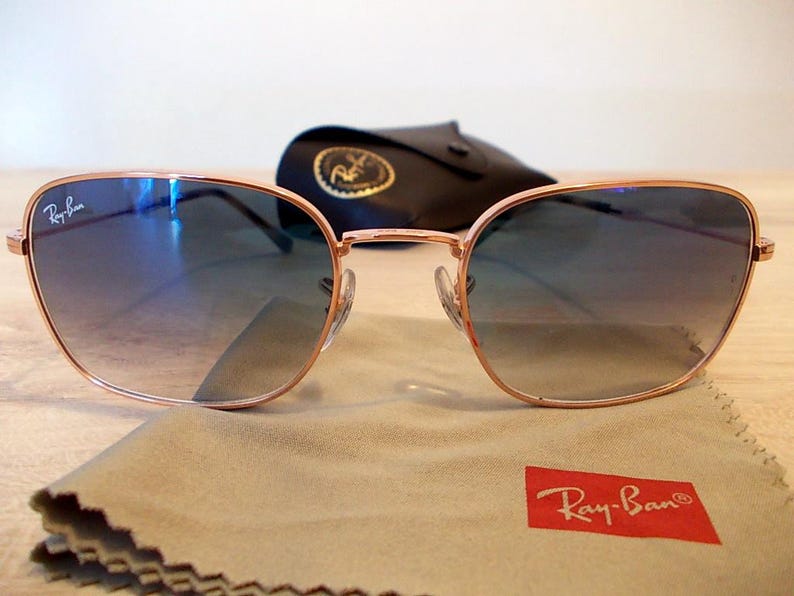 Pu&ograve; includere: Occhiali da sole con montatura color oro rosa e lenti blu sfumate. Il logo Ray-Ban &egrave; visibile sulla lente superiore sinistra e su un rettangolo rosso sul panno per la pulizia. Sullo sfondo, una custodia nera con un'emblema dorato.