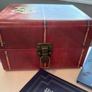 Puede incluir: Una caja roja con un escudo de Hogwarts en la tapa, con un cierre de latón y detalles decorativos. La caja está acompañada de dos libros con cubiertas azules y blancas. La caja está diseñada para parecer un baúl vintage.