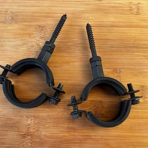 May include: Two black pipe clamps with screws and bolts are shown on a wooden surface. The clamps are circular with a split design, allowing them to encircle a pipe. The screws are threaded and extend upwards.