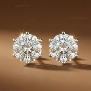 May include: A pair of sparkling diamond stud earrings. Each earring features a large, round-cut, clear gemstone set in a silver-colored metal. The earrings are displayed on a brown background, with light reflections.