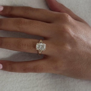 May include: A gold ring featuring a cushion-cut centre stone surrounded by a milgrain-detailed bezel setting. The band has a split-shank design that tapers towards the centre stone, creating a structured look with a polished metallic finish.