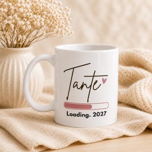 Könnte beinhalten: Weiße Keramik-Tasse mit dem Wort "Tante" in Handschrift, zwei kleinen rosa Herzen und einer Ladebalken-Grafik mit dem Text "Loading. 2027". Die Tasse hat einen gebogenen Henkel.