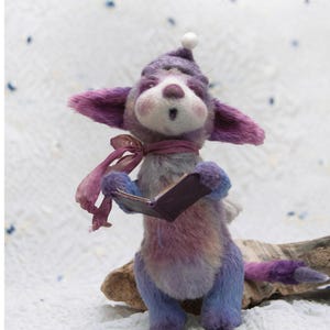 May include: A purple and blue plush toy animal wearing a white hat and a pink scarf, holding a small book. The toy is standing on a piece of driftwood.