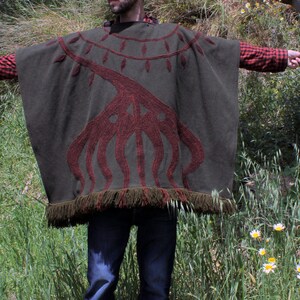 May include: A green poncho with a red embroidered design of a tree with branches. The poncho has a fringe border.