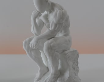 May include: A matte white figurine depicting a muscular human figure in a seated, contemplative pose with one hand resting against the chin. The figure is perched upon a textured, rock-like base, showcasing detailed anatomical muscle definition.
