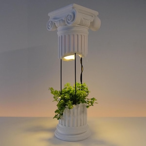 Può includere: Un vaso bianco a forma di colonna classica spezzata con base e capitello scanalati. Le due sezioni sono collegate da sottili aste di metallo nero, con una piccola luce integrata nella parte inferiore del capitello che illumina una pianta verde.