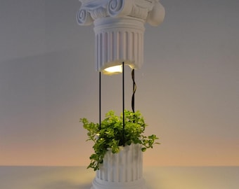 May include: A white planter shaped like a broken classical column with a fluted base and capital. The two sections are connected by thin black metal rods, with a small light integrated into the underside of the capital, illuminating a green plant.